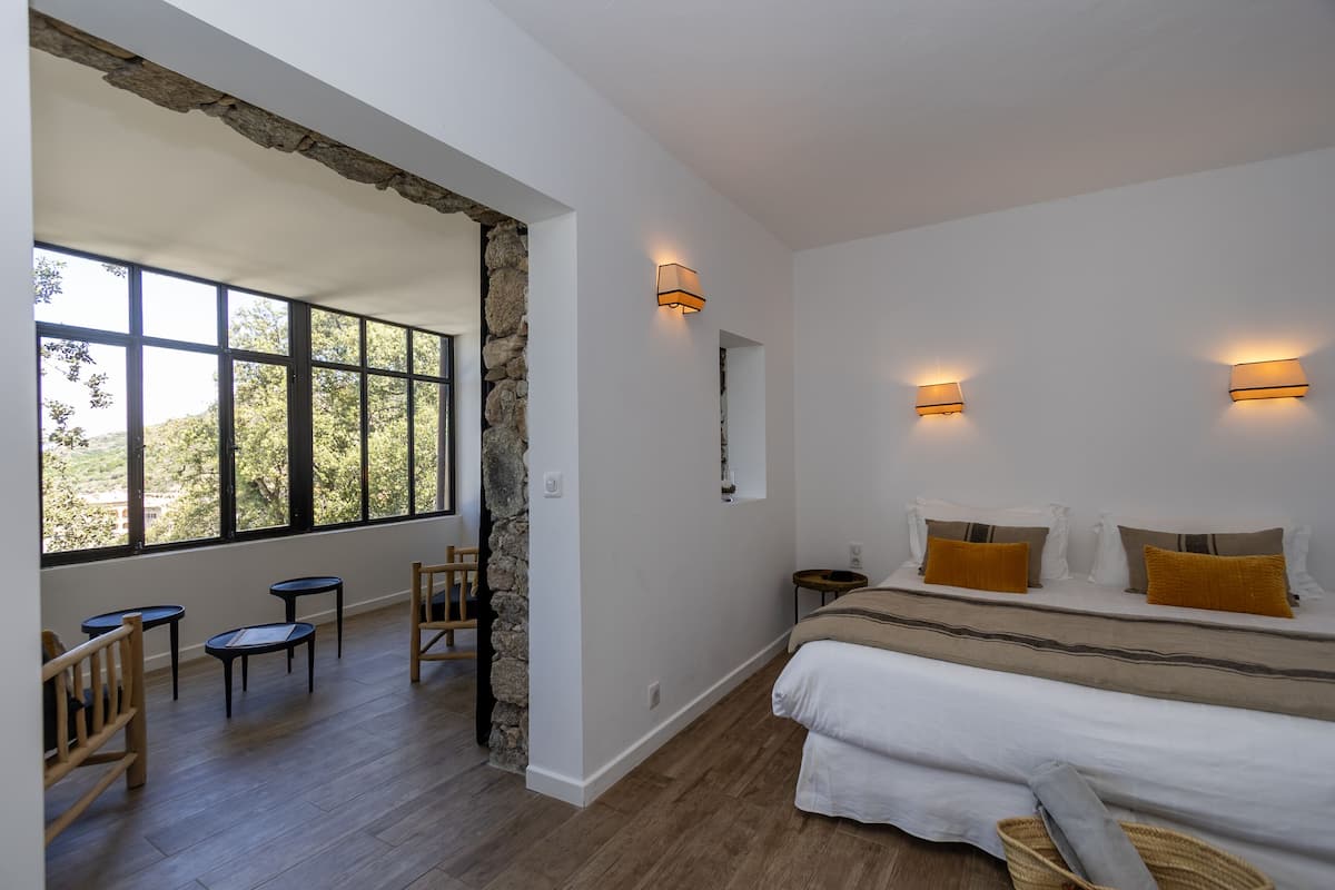 A bedroom with a large window, a bed with white and mustard pillows, a stone accent wall, and two wall-mounted lights.