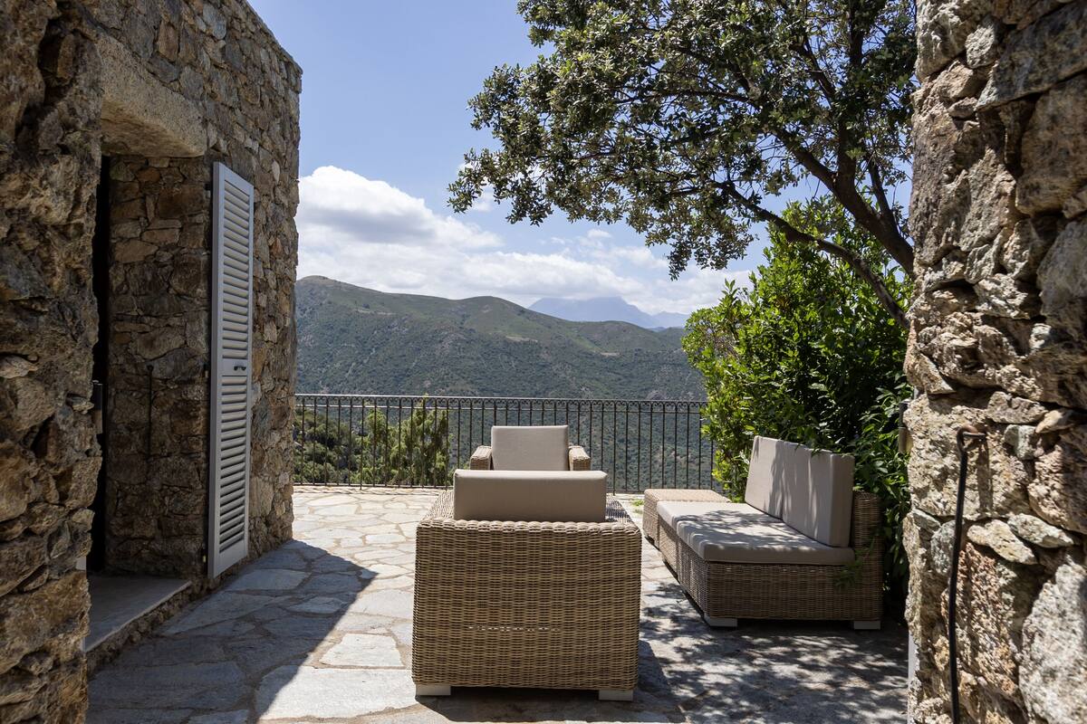 A stone-walled outdoor seating area with wicker furniture and a view of mountains.