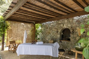 A room with a bed, a wooden chair, a small table with a vase, and a stone wall with an arched window. - Hôtel Case Latine (Lama)