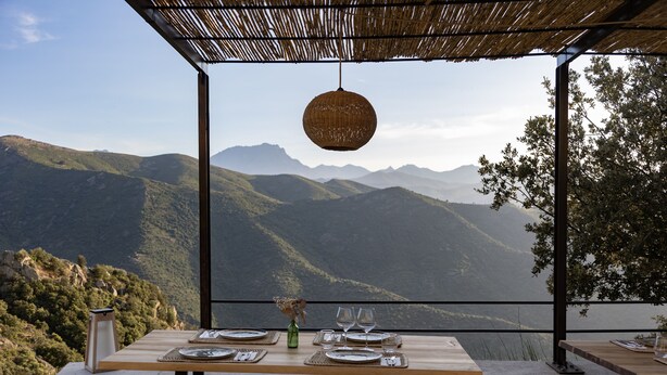 Una mesa de comedor dispuesta con platos, vasos y un jarrón, con vistas a un paisaje montañoso.