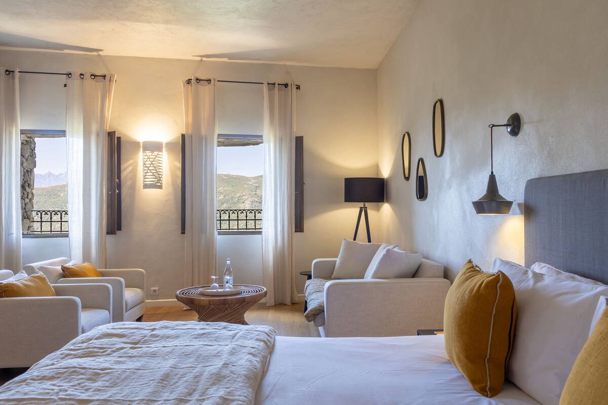 A hotel room with a bed, two armchairs, a sofa, a small table, and a view of the mountains through the windows.