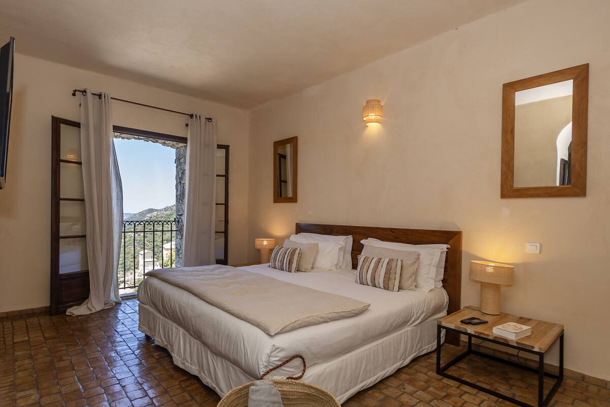 A bedroom with a large bed, a nightstand, a lamp, and a view of the outdoors through a window with curtains.