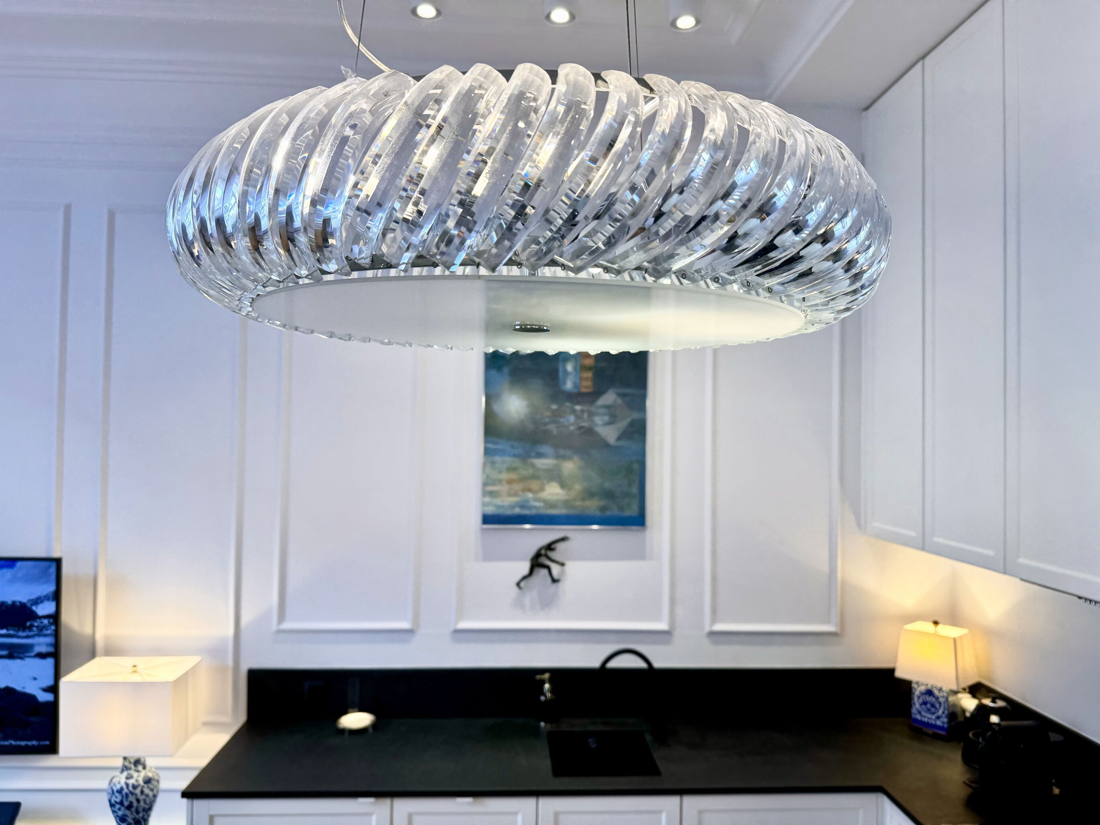 A modern kitchen with a unique, ribbed pendant light, a black countertop, white cabinetry, and a framed artwork above the sink.