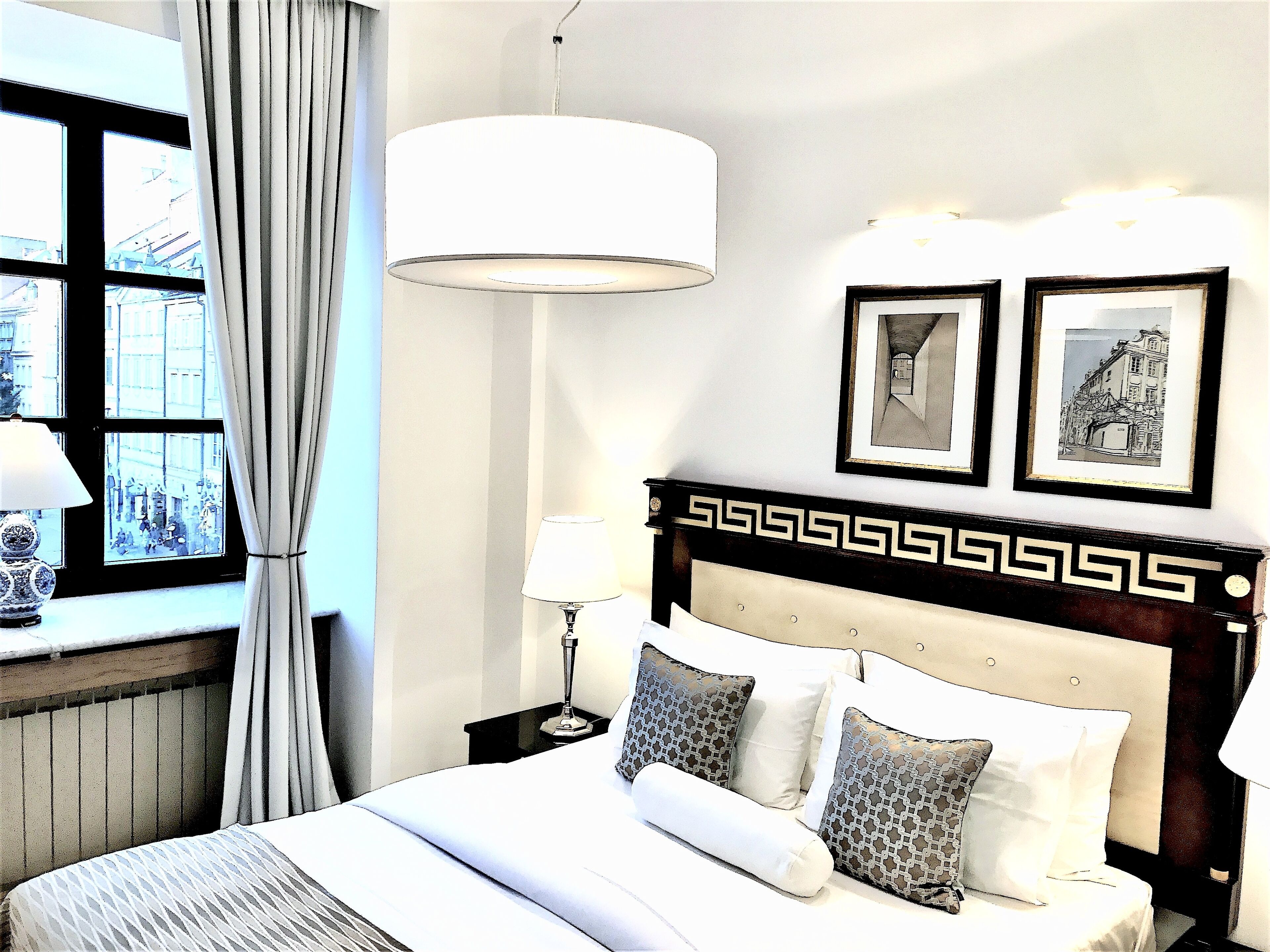 Une chambre d’hôtel avec un grand lit, une table de chevet avec une lampe, une fenêtre avec des rideaux et deux tableaux encadrés au mur.