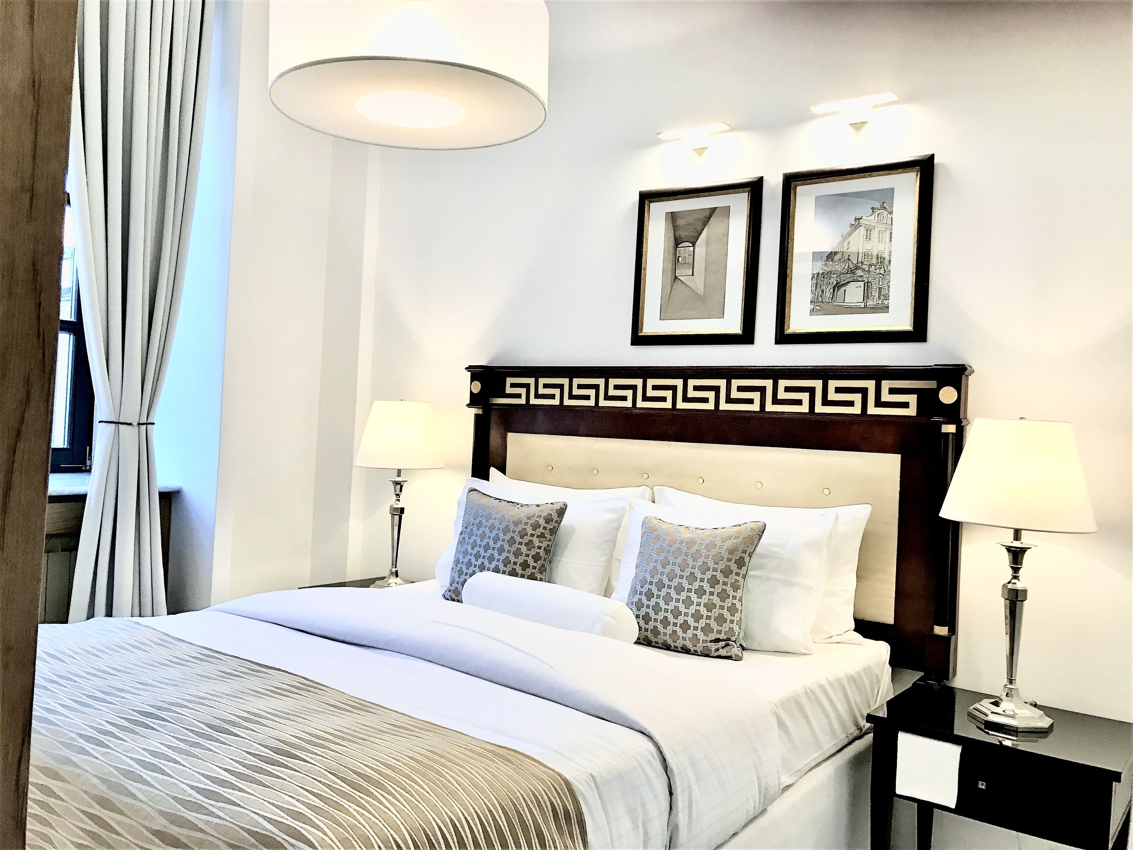 A neatly made bed with a patterned bedspread, two bedside lamps, and a headboard with a decorative design. There are two framed pictures on the wall above the bed.