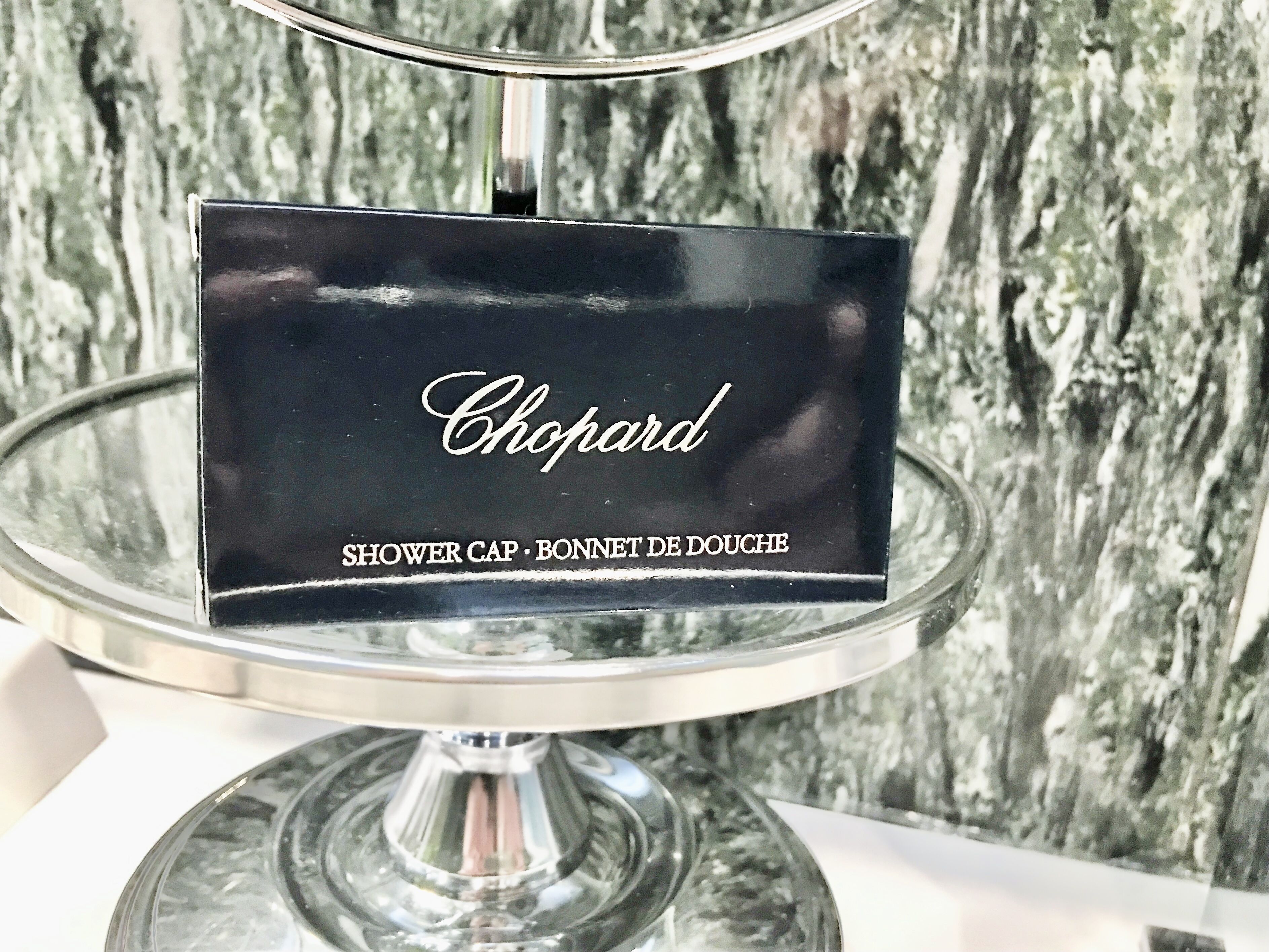 Een zwart bord met de tekst 'Chopard' en 'SHOWER CAP - BONNET DE DOUCHE' op een transparante glazen standaard, voor een structuurwand.