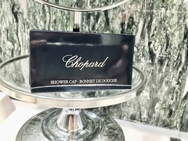 A black sign with the word "Chopard" and the text "SHOWER CAP - BONNET DE DOUCHE" on a clear glass stand against a textured wall.
