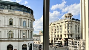 Vista de un edificio histórico con arquitectura clásica, que incluye columnas y ventanas arqueadas, desde una ventana con vista a una calle con peatones y coches estacionados.