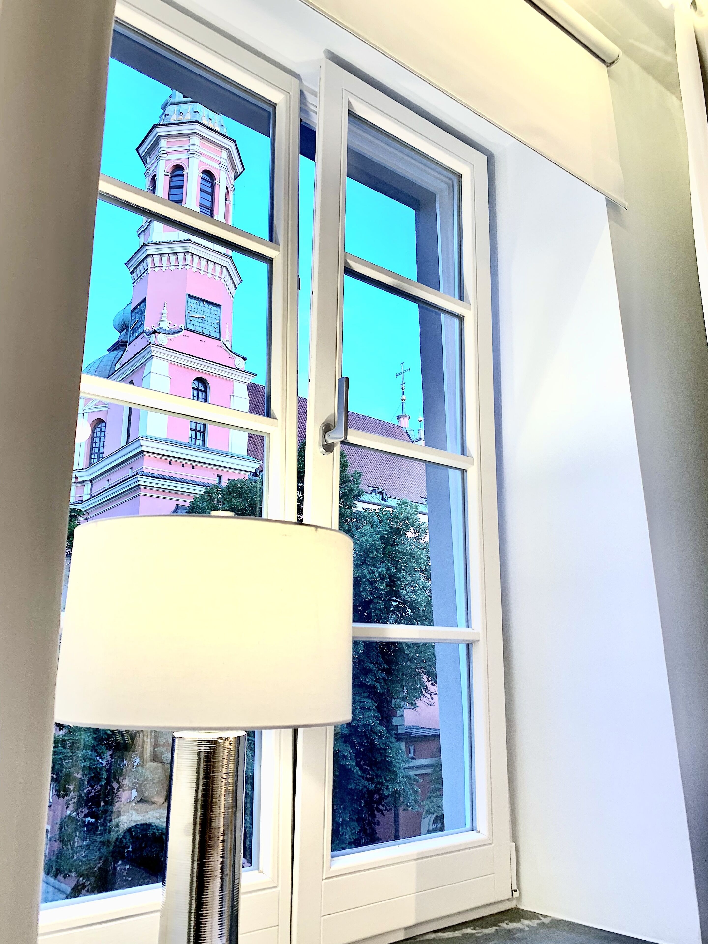 Janela com vista para um edifício rosa com torre do relógio, uma parede branca e uma persiana.