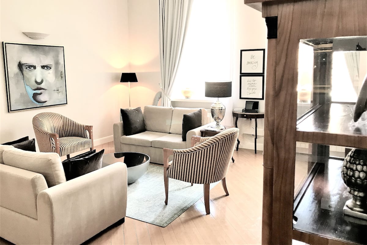 Una sala de estar moderna con un sofá, sillones y una mesa de centro. Hay un cuadro en la pared, una lámpara y un ventanal con cortinas.