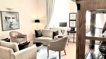 A modern living room with a sofa, armchairs, and a coffee table. There is a painting on the wall, a lamp, and a window with curtains.