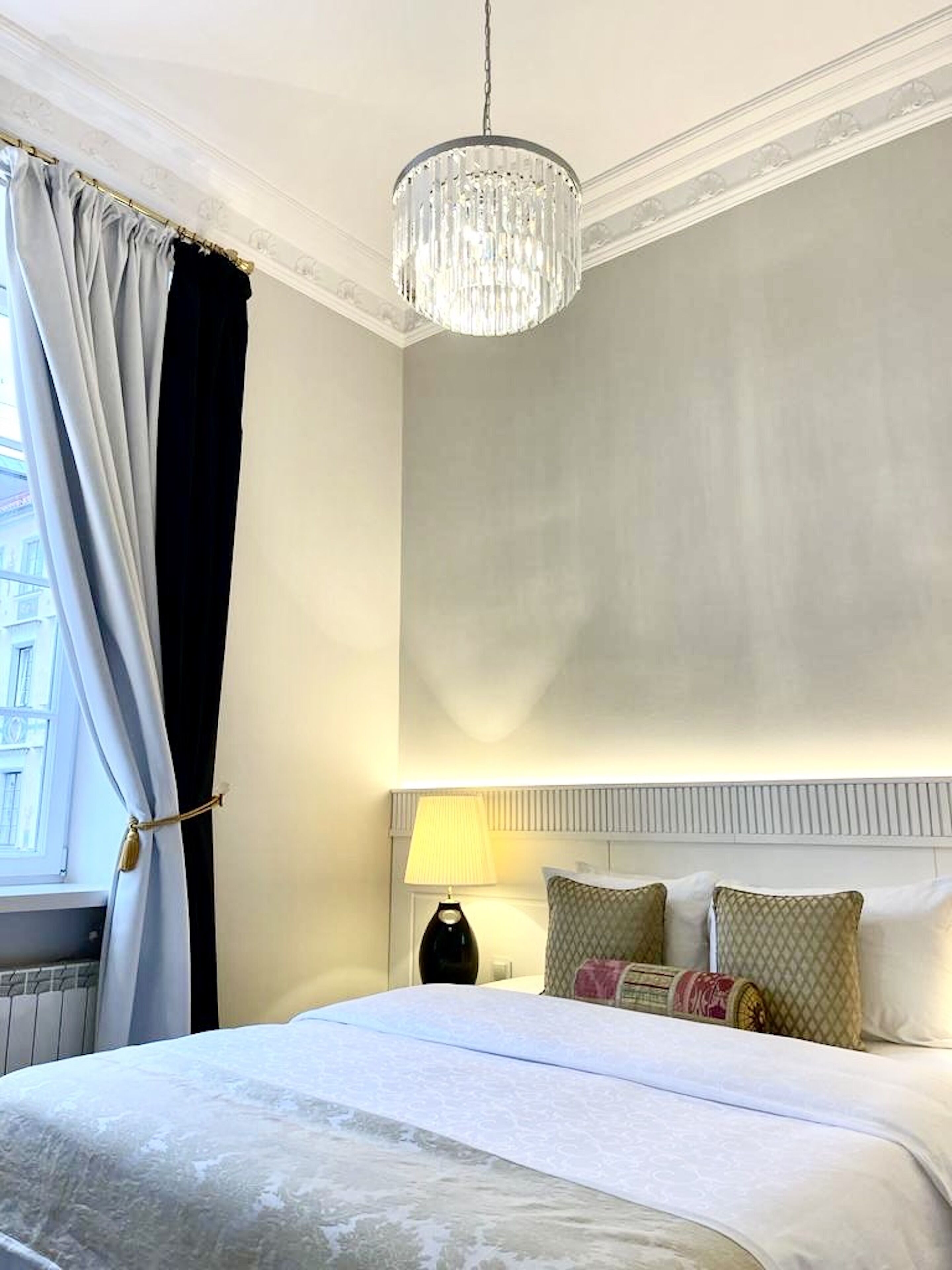 A hotel room with a large bed, a chandelier, a nightstand with a lamp, and a window with curtains.