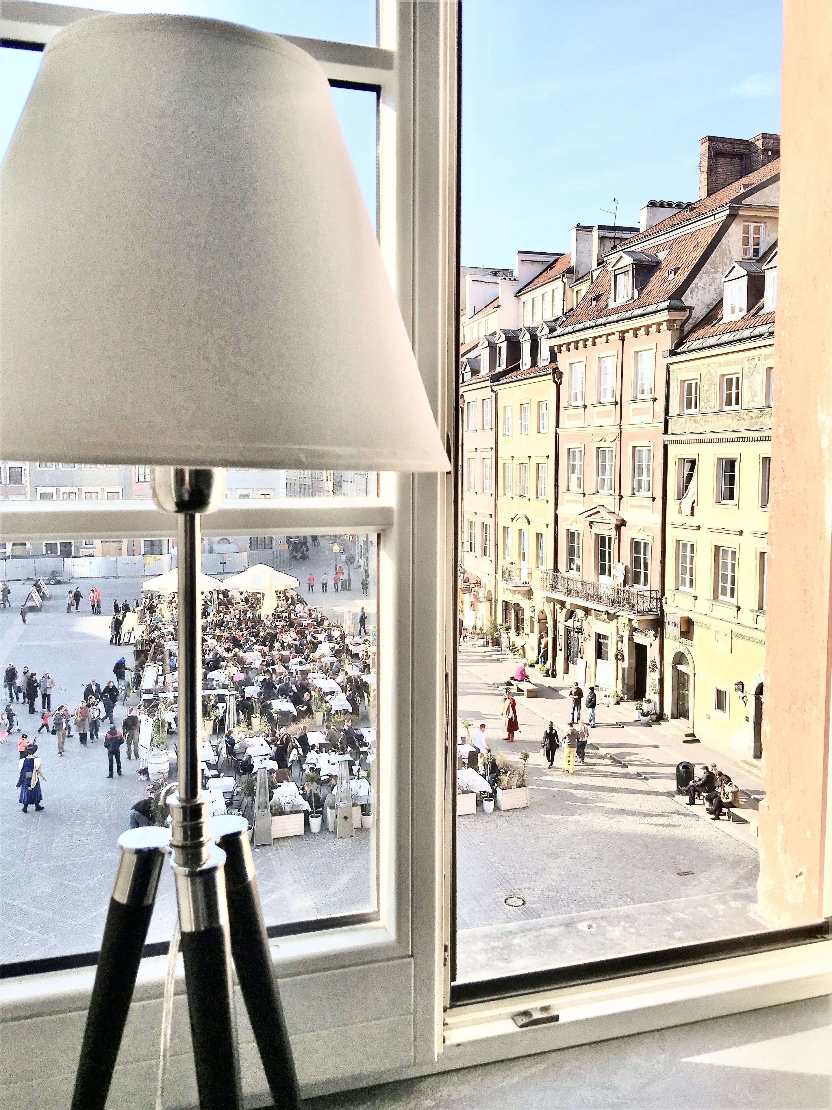 Ein Fenster mit einer Lampe, das auf einen belebten Platz mit Gebäuden im Hintergrund blickt.