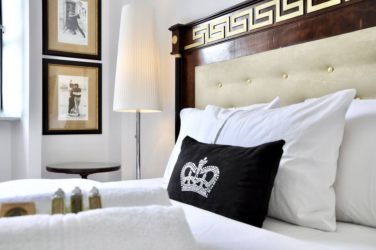 Habitación de hotel con una cama, cabecera con motivo decorativo, cuadros en la pared y mesita de noche con lámpara.