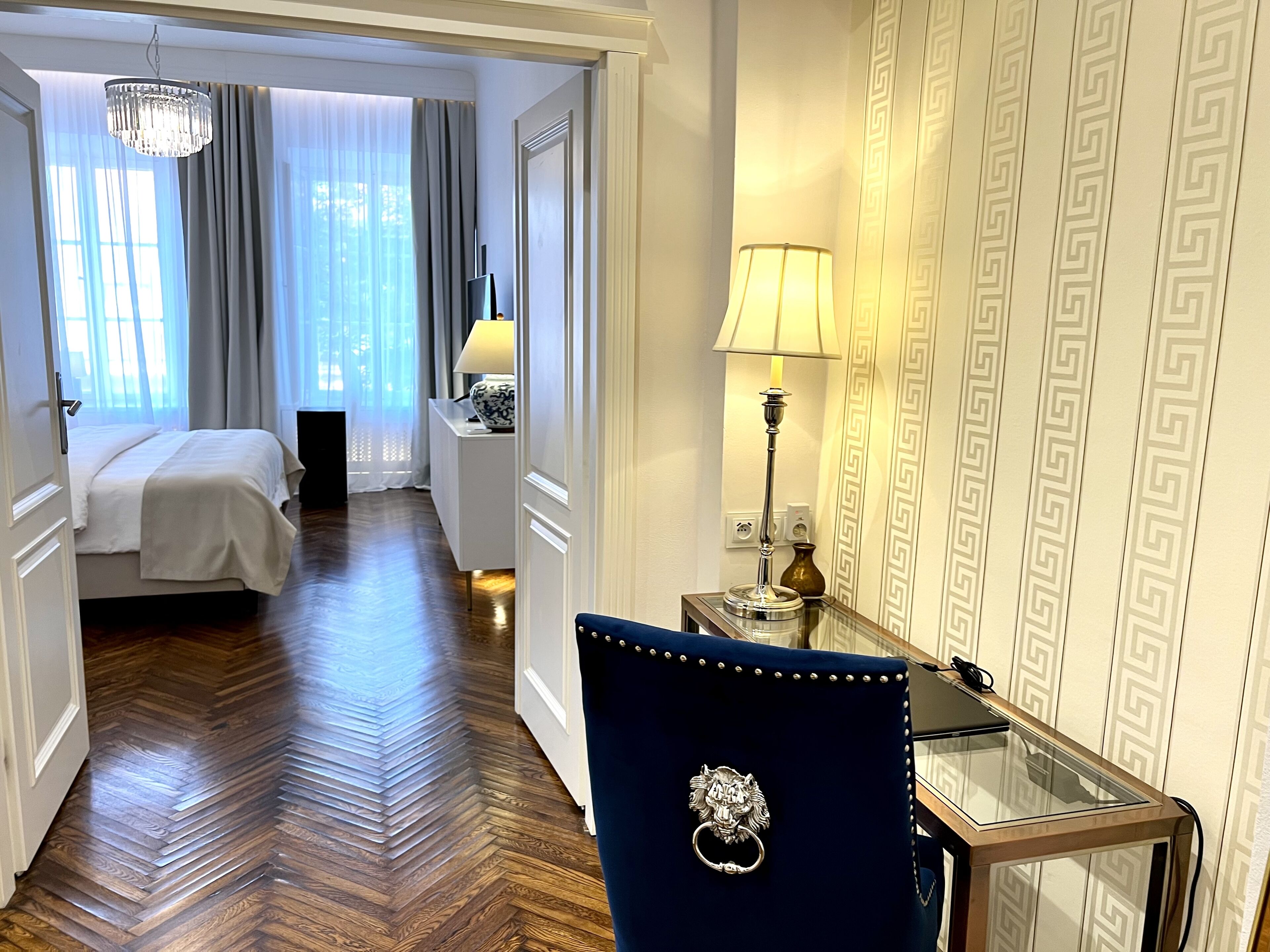 Une chambre d’hôtel avec un lit, un bureau doté d’une lampe, une chaise et un couloir menant à une autre pièce.