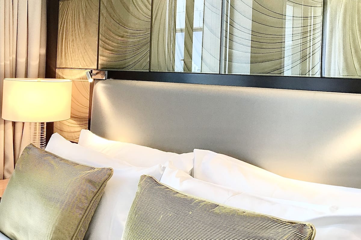 Una habitación de hotel con un ventanal grande, una cama con ropa blanca y una lámpara en una mesita de noche.