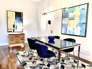 Uma sala de jantar com mesa de vidro, cadeiras azuis, tapete estampado e quadros abstratos nas paredes.