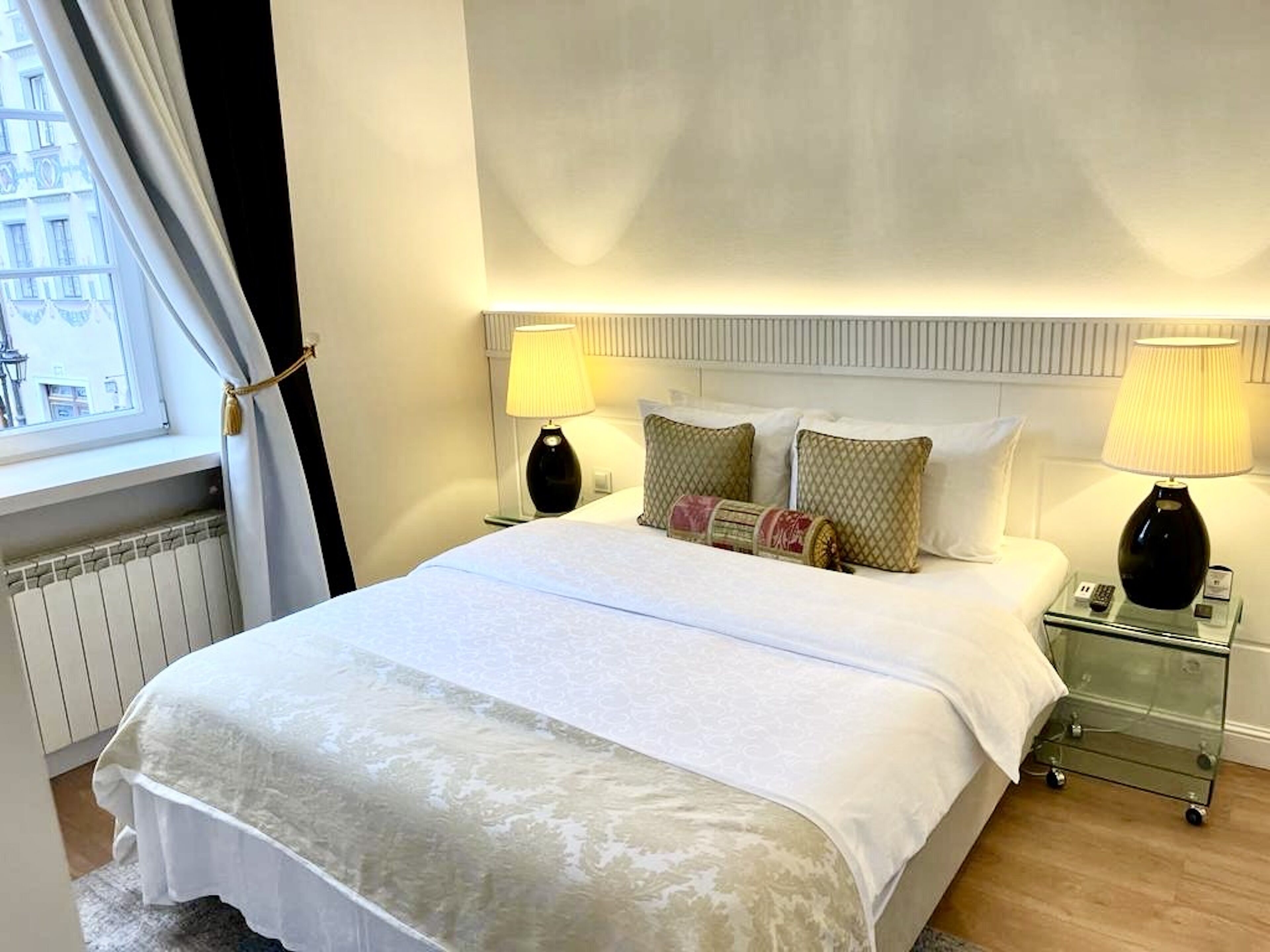 A neatly made bed with a white duvet, two bedside lamps, and a small bedside table with a phone and a lamp.