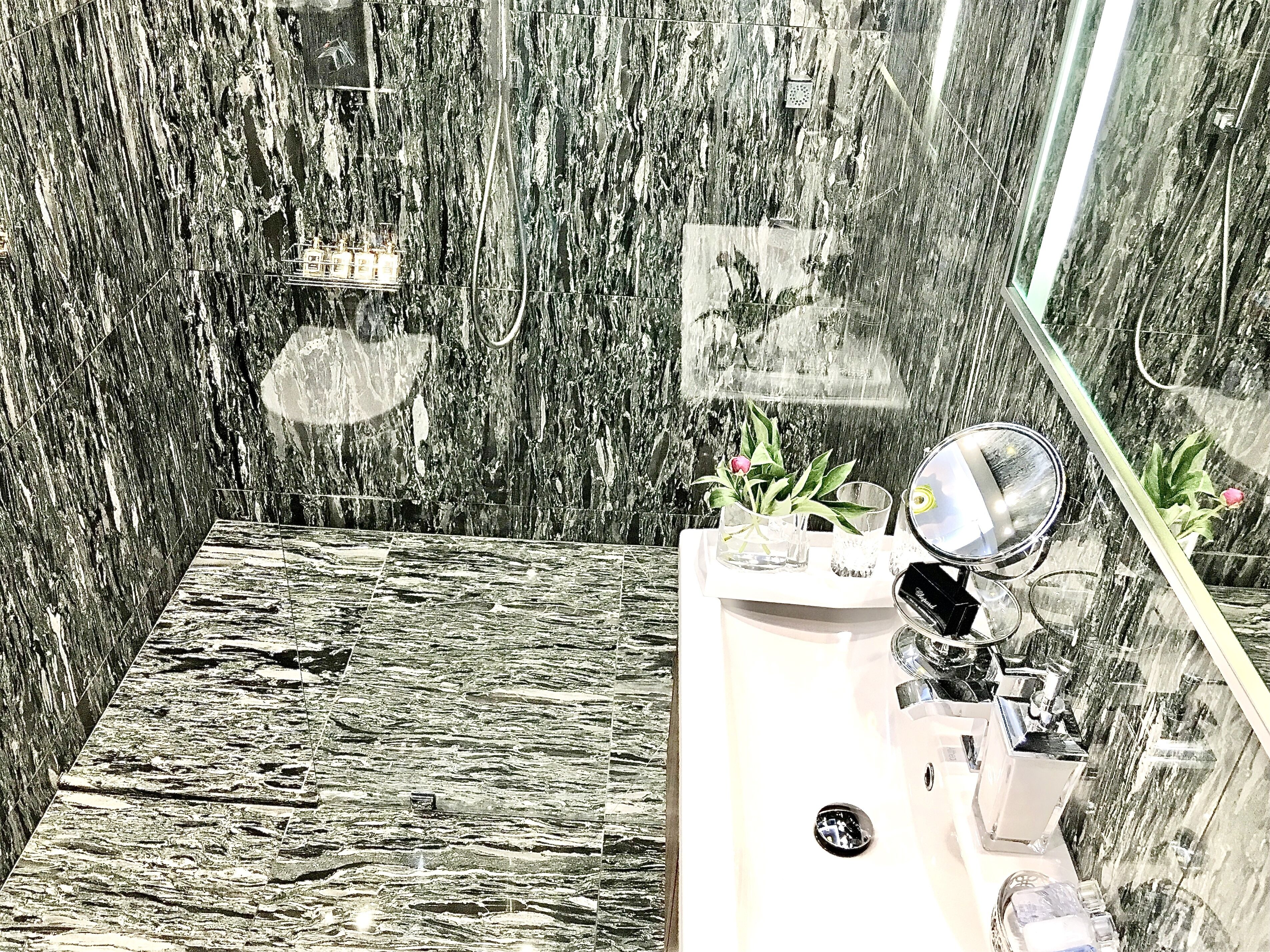A bathroom with a marble wall and floor, a sink with a mirror, and a small plant.