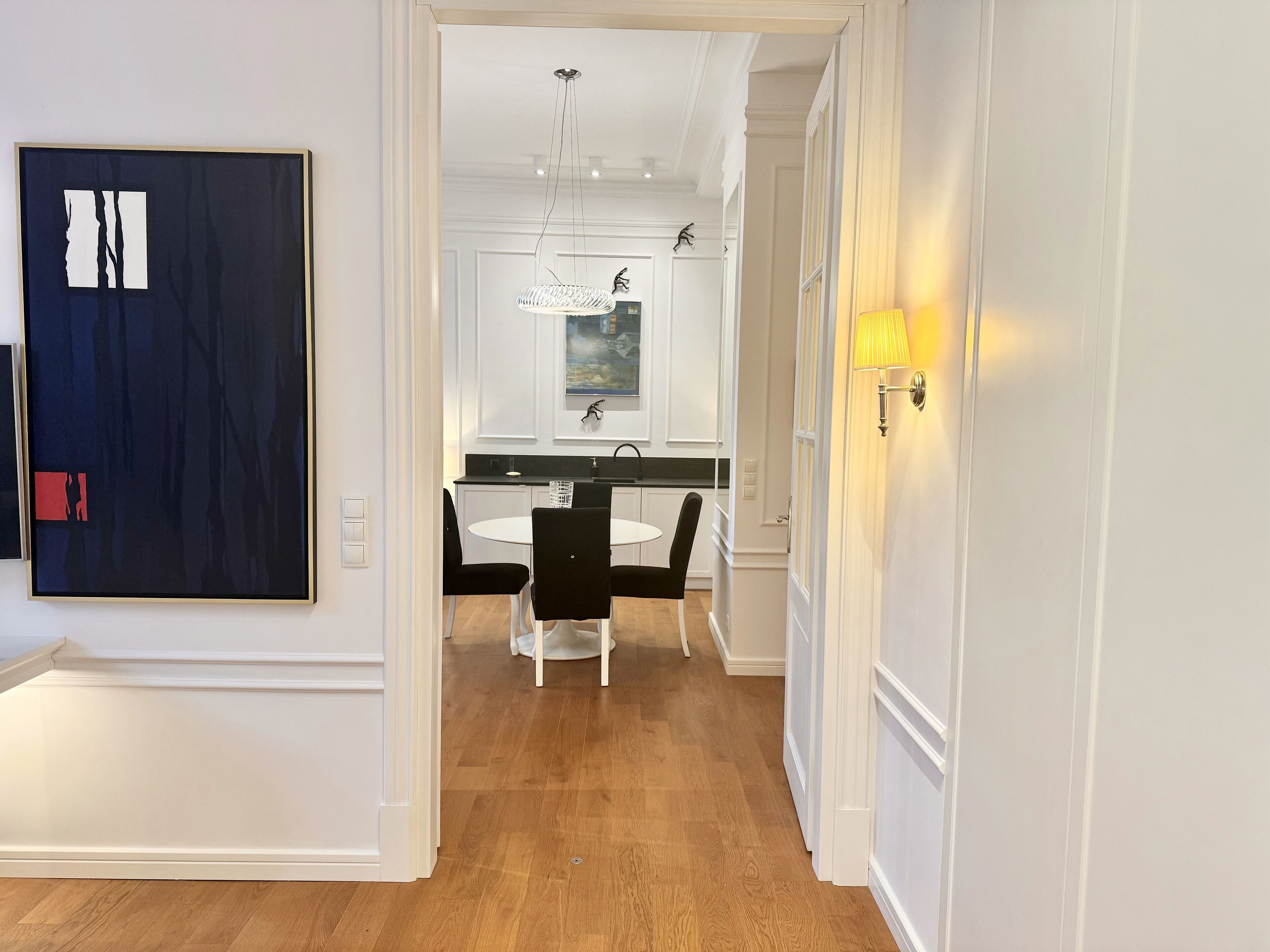 Un couloir mène à une salle à manger où se trouvent une table et des chaises blanches, un luminaire suspendu et une œuvre d’art encadrée au mur.