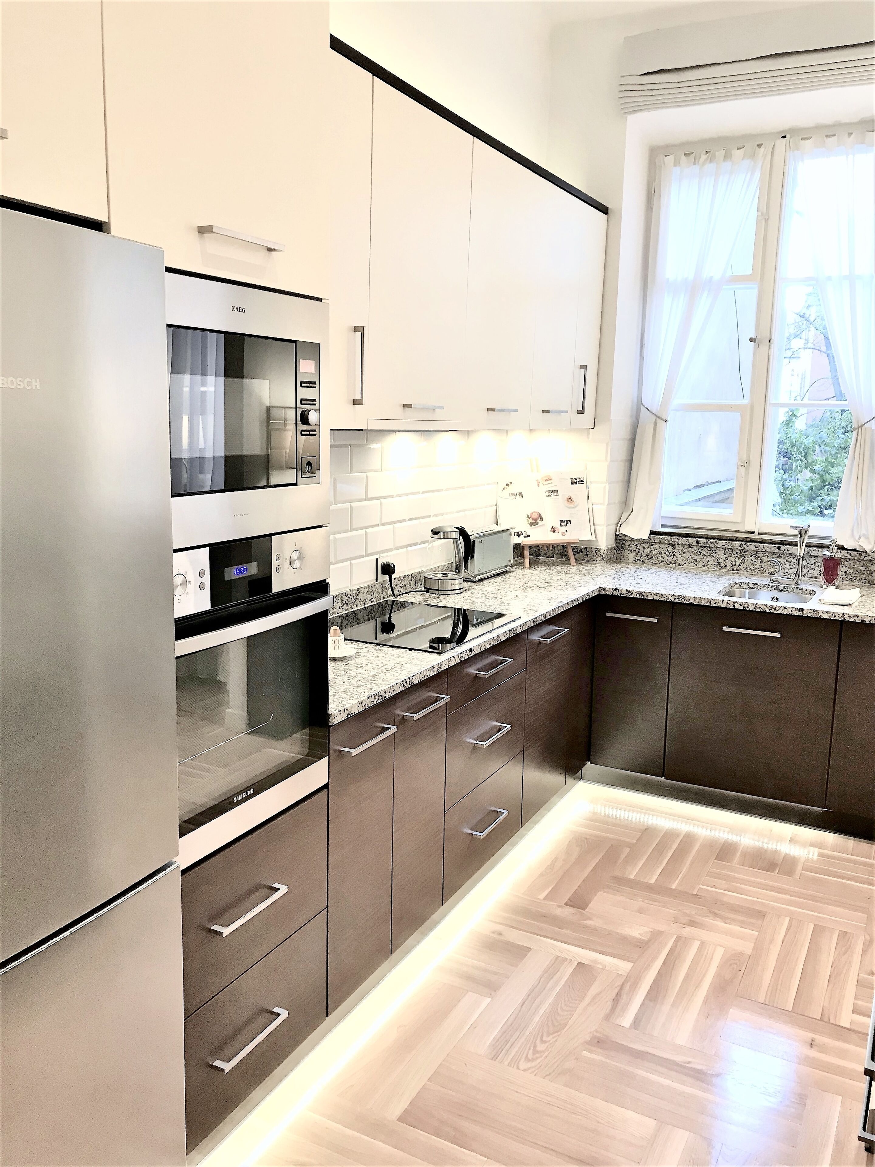 A modern kitchen with stainless steel appliances, a herringbone wood floor, and a window with curtains.