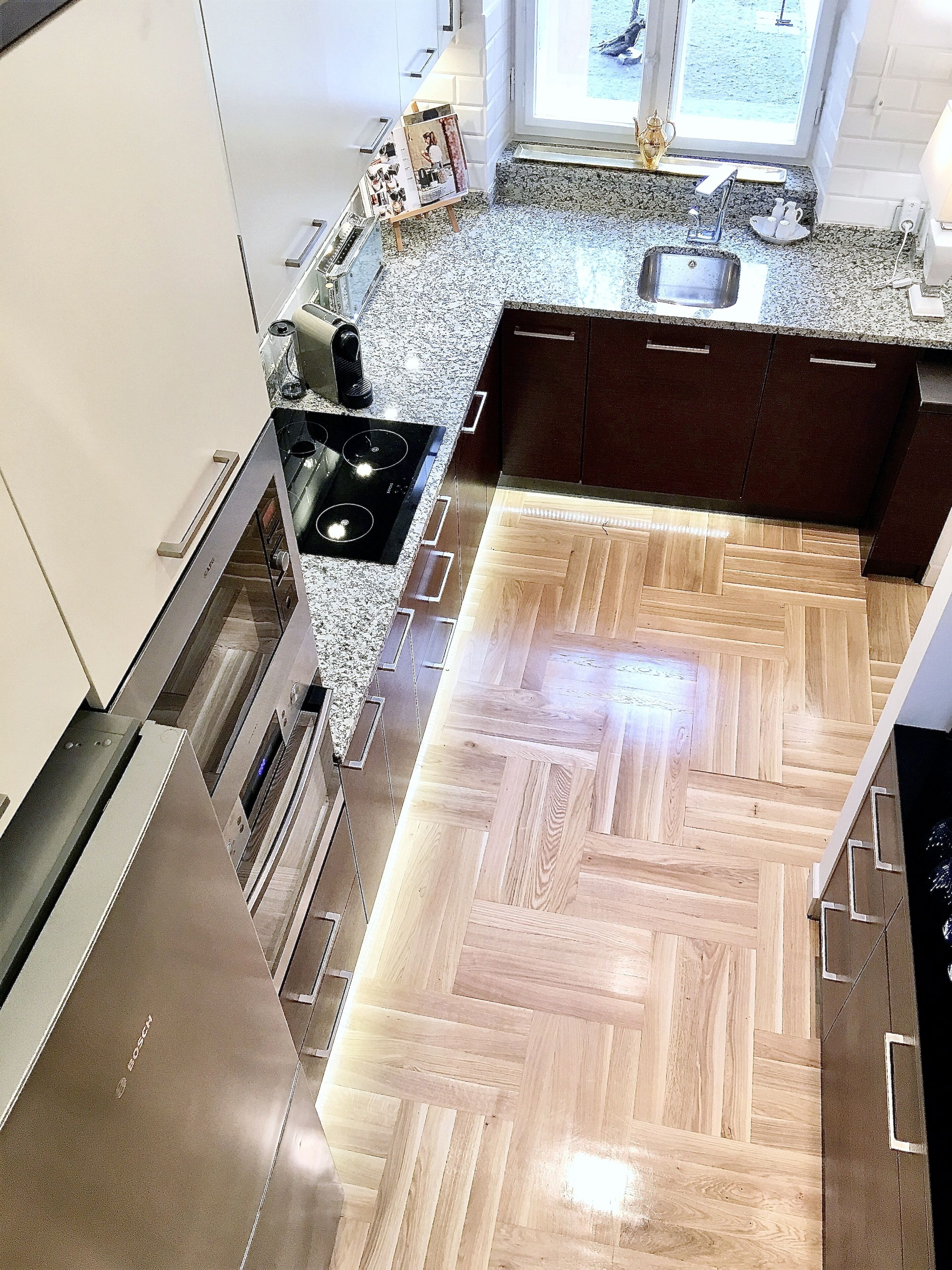 Una cocina moderna con piso de madera en patrón de espiga, electrodomésticos de acero inoxidable y encimera de granito.