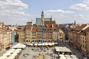 Une place historique, dominée par un bâtiment remarquable avec une tour, et offrant un vaste espace ouvert où se trouvent des étals de marché et des parasols.