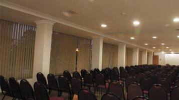 Uma sala de conferências com fileiras de cadeiras, palco e uma grande janela com persianas.
