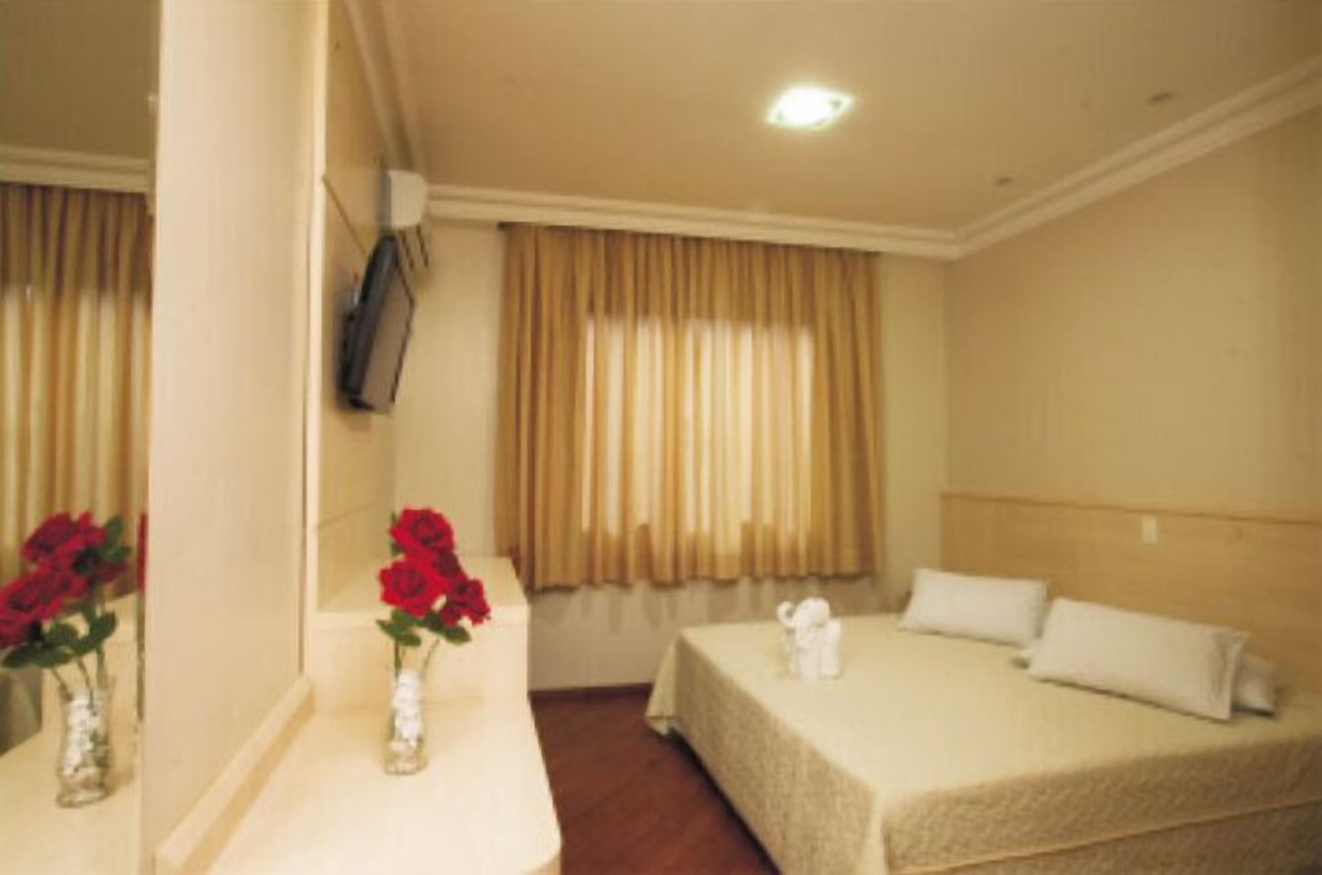 Habitación de hotel con cama, mesita de noche con un florero de flores rojas, espejo y ventana con cortinas.