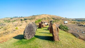 A grassy area with large rocks and a small building in the background. - Nisarg Resort (Madale)