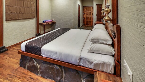 A bedroom with a wooden bed, stone foundation, and a mirror reflecting a wooden wardrobe and a door. - Nisarg Resort (Madale)