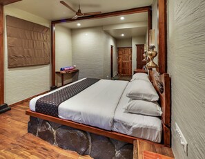 A bedroom with a wooden bed, stone foundation, and a mirror reflecting a wooden wardrobe and a door. - Nisarg Resort (Madale)