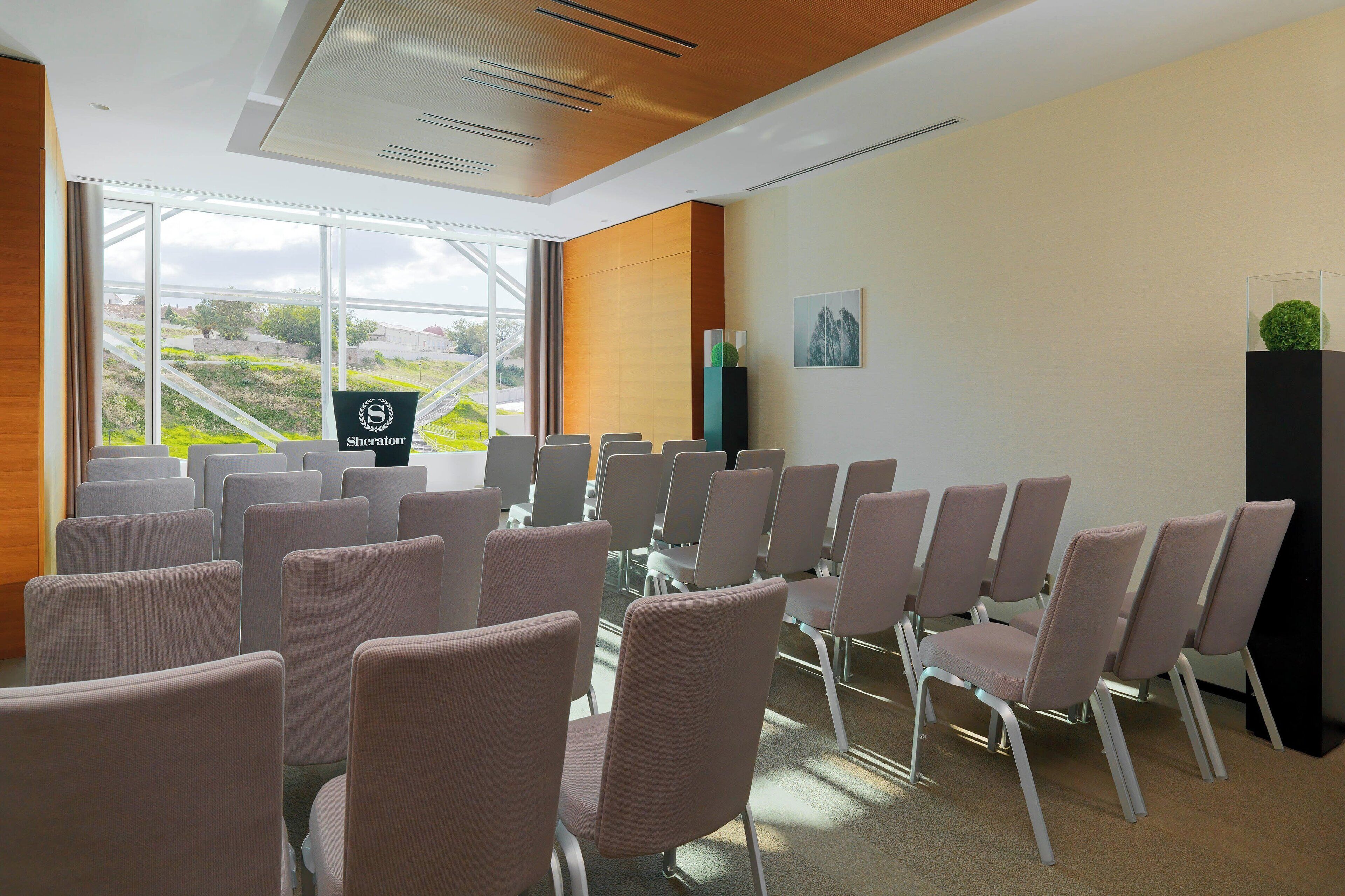 Un salón de conferencias con filas de sillas, el logotipo de Sheraton y vistas al exterior con vegetación.