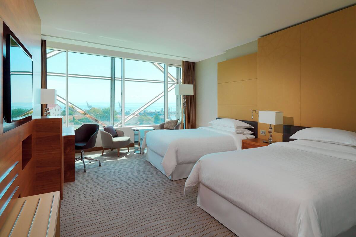 Ein Hotelzimmer mit zwei Betten, einem Schreibtisch, einem Stuhl und einem großen Fenster mit Blick auf die Stadt.