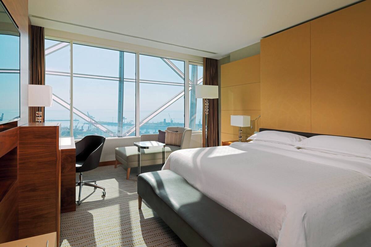 Ein Hotelzimmer mit einem großen Bett, einem Schreibtisch, einem Sessel, einem kleinen Tisch und Blick auf die Stadt durch große Fenster.
