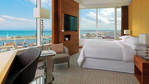 A hotel room with a large bed, a desk with a chair, a TV mounted on the wall, and a view of the sea and cityscape. - Sheraton Annaba Hotel (Annaba)