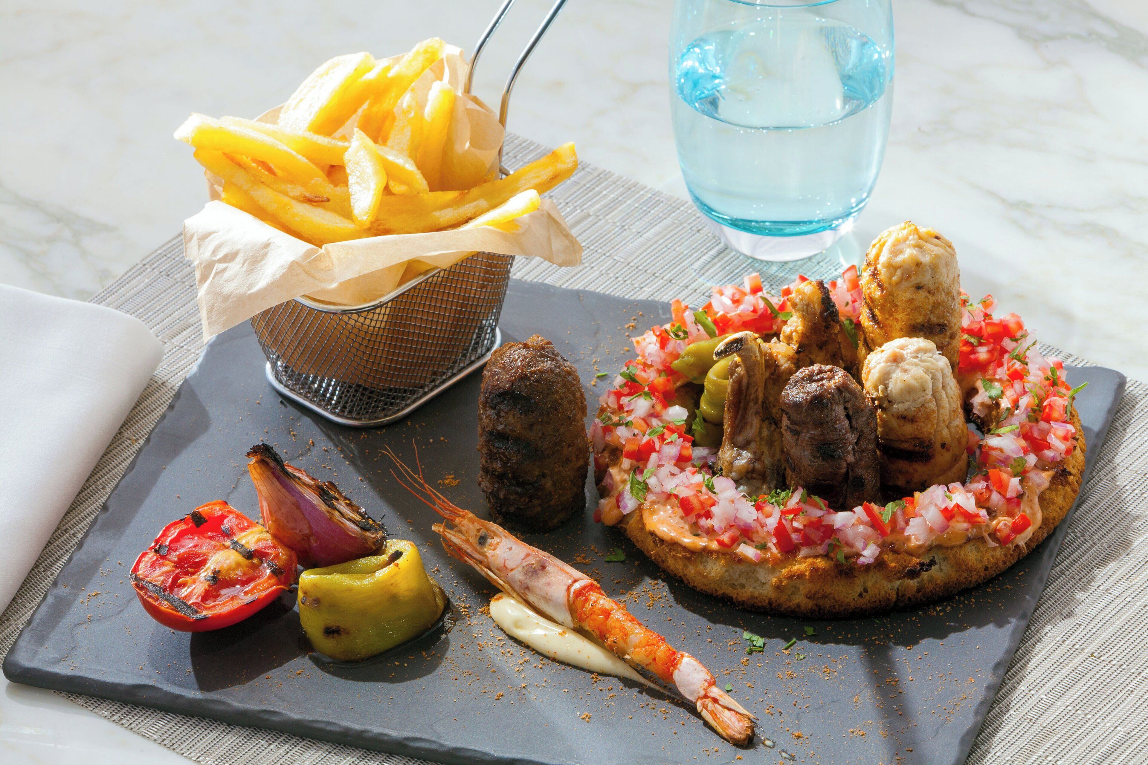 A meal consisting of grilled meat, shrimp, and a vegetable dish, served on a slate plate with a side of fries and a glass of water.