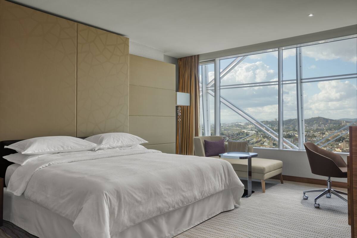 Ein Hotelzimmer mit einem großen Bett, einem kleinen Tisch, einem Sessel und Blick auf eine Stadtlandschaft durch ein großes Fenster.