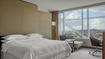 A hotel room with a large bed, a small table, a chair, and a view of a cityscape through a large window.