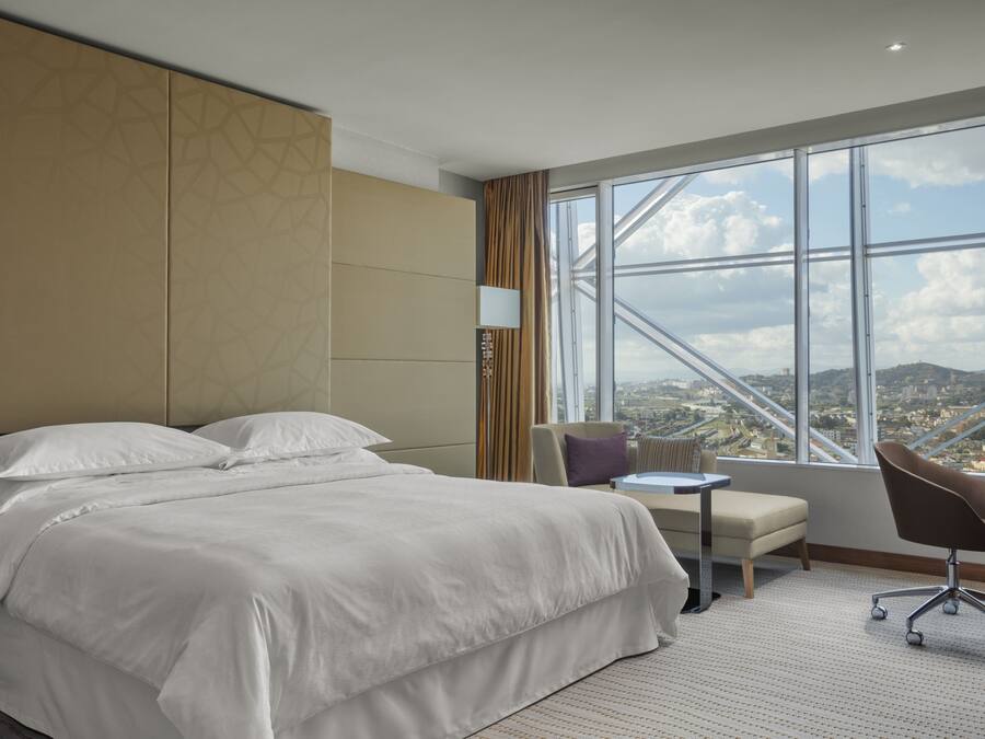 A hotel room with a large bed, a small table, a chair, and a view of a cityscape through a large window.