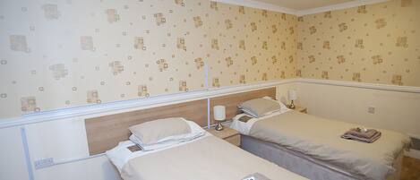 A hotel room with two single beds, a wooden headboard, a nightstand with a lamp, and patterned wallpaper.