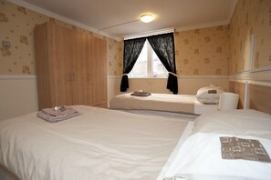 A hotel room with two beds, a wardrobe, a window with curtains, and a bedside table with a lamp.