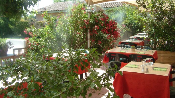 An outdoor dining area with tables covered in red tablecloths, surrounded by lush greenery and flowering plants. - Hotel Saint Louis (Le Thor)