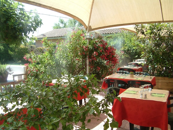 An outdoor dining area with tables covered in red tablecloths, surrounded by lush greenery and flowering plants. - Hotel Saint Louis (Le Thor)