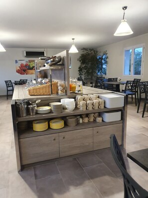 A buffet setup with various dishes and utensils, a plant, and seating arrangements in a well-lit room. - Hotel Saint Louis (Le Thor)