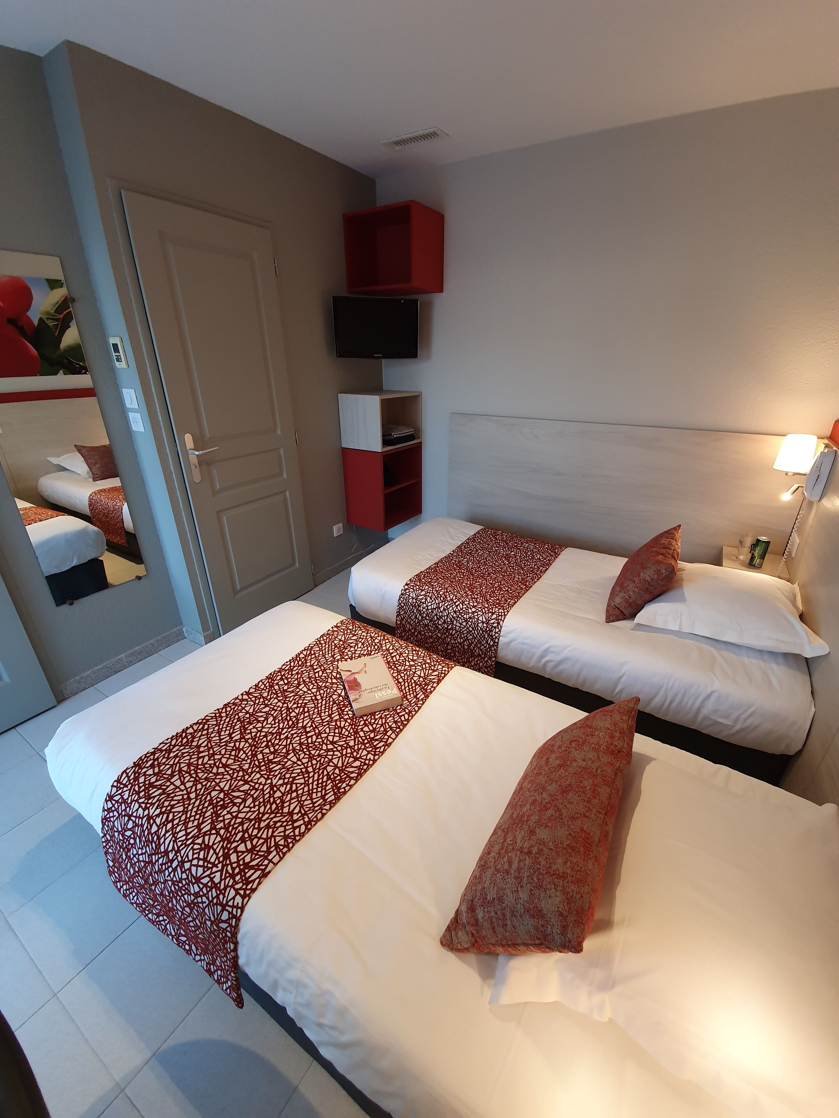 A hotel room with two beds, a TV mounted on the wall, a bedside table with a lamp, and a red storage unit.