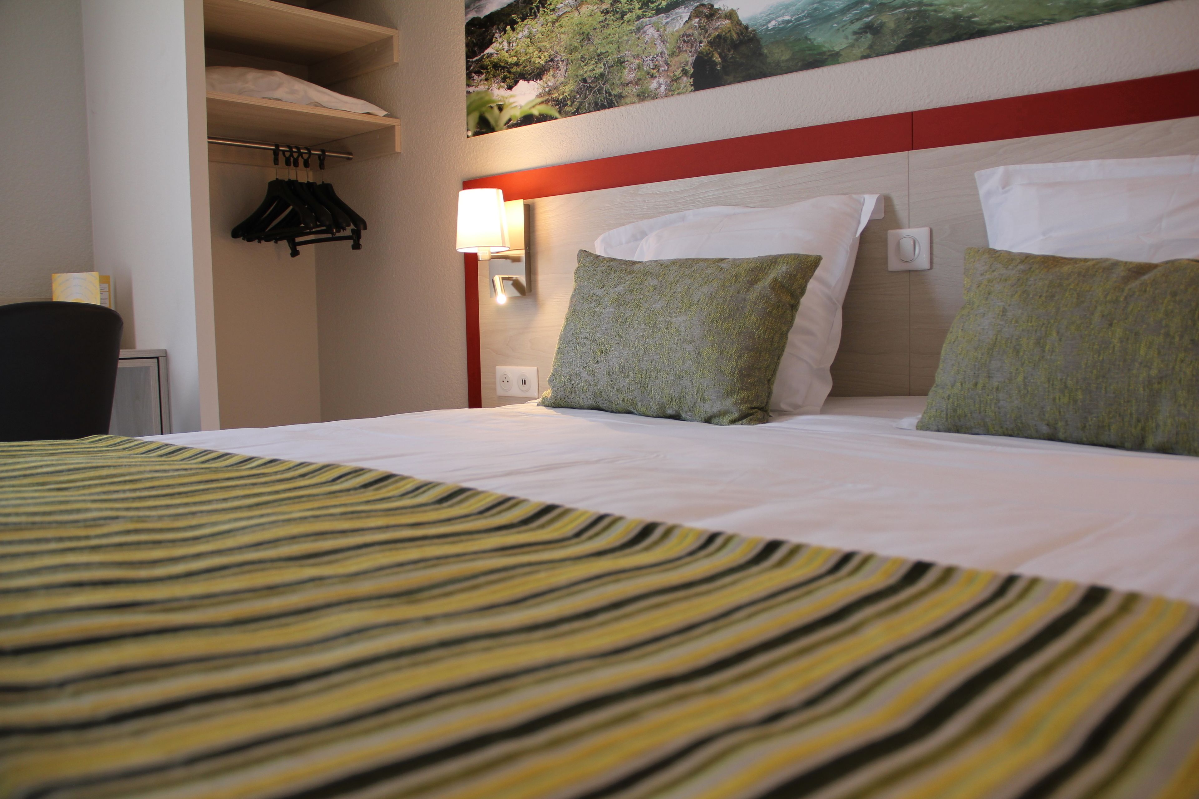 A hotel room with a bed, a nightstand with a lamp, a coat rack, and a painting on the wall.