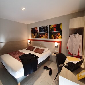 A hotel room with a bed, a desk with a laptop, a chair, and a coat rack with a shirt. - Hotel Saint Louis (Le Thor)