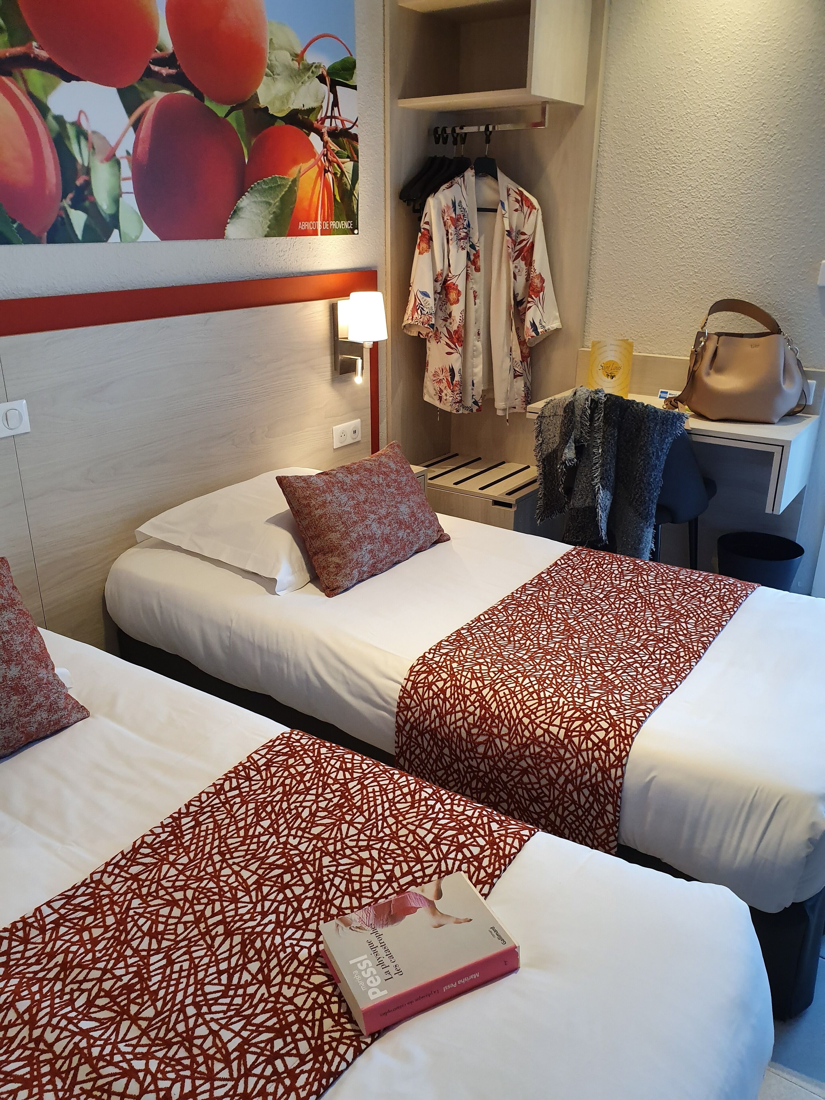 Habitación de hotel con dos camas, una mesita de noche, una lámpara, un perchero y un cuadro con motivos de frutas.