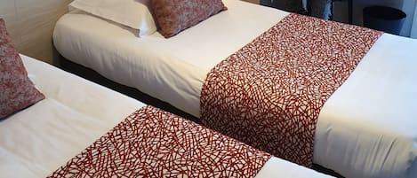 A hotel room with two beds, a nightstand, a lamp, a coat rack, and a wall with a fruit-themed painting.